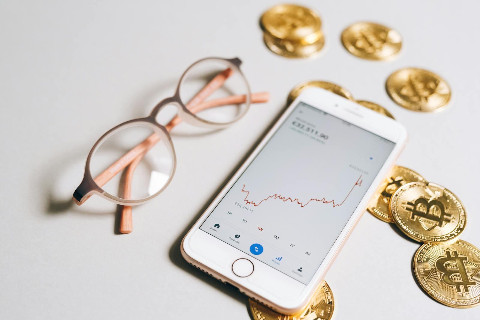 Smartphone showing a cryptocurrency price chart beside physical Bitcoin coins.