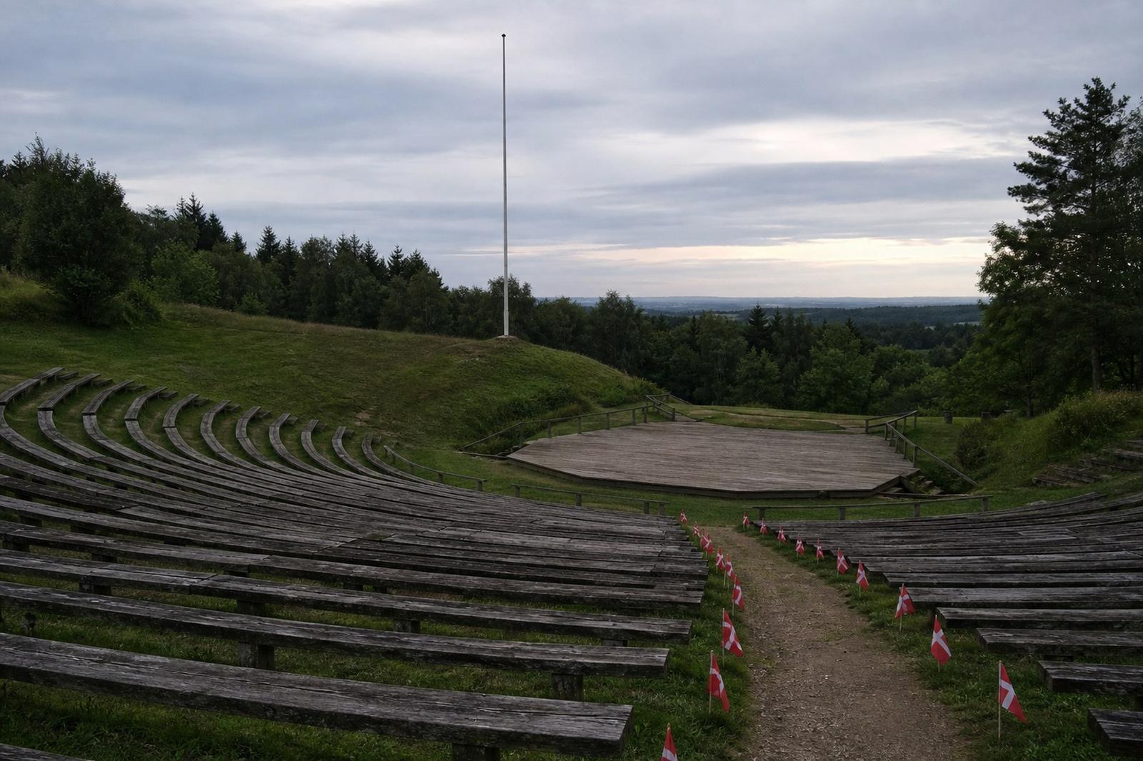 Empty wooden benches in the grassy Rebild hills amphitheatre in Denmark, with small Danish flags along a path and a bare flagpole against an overcast sky.