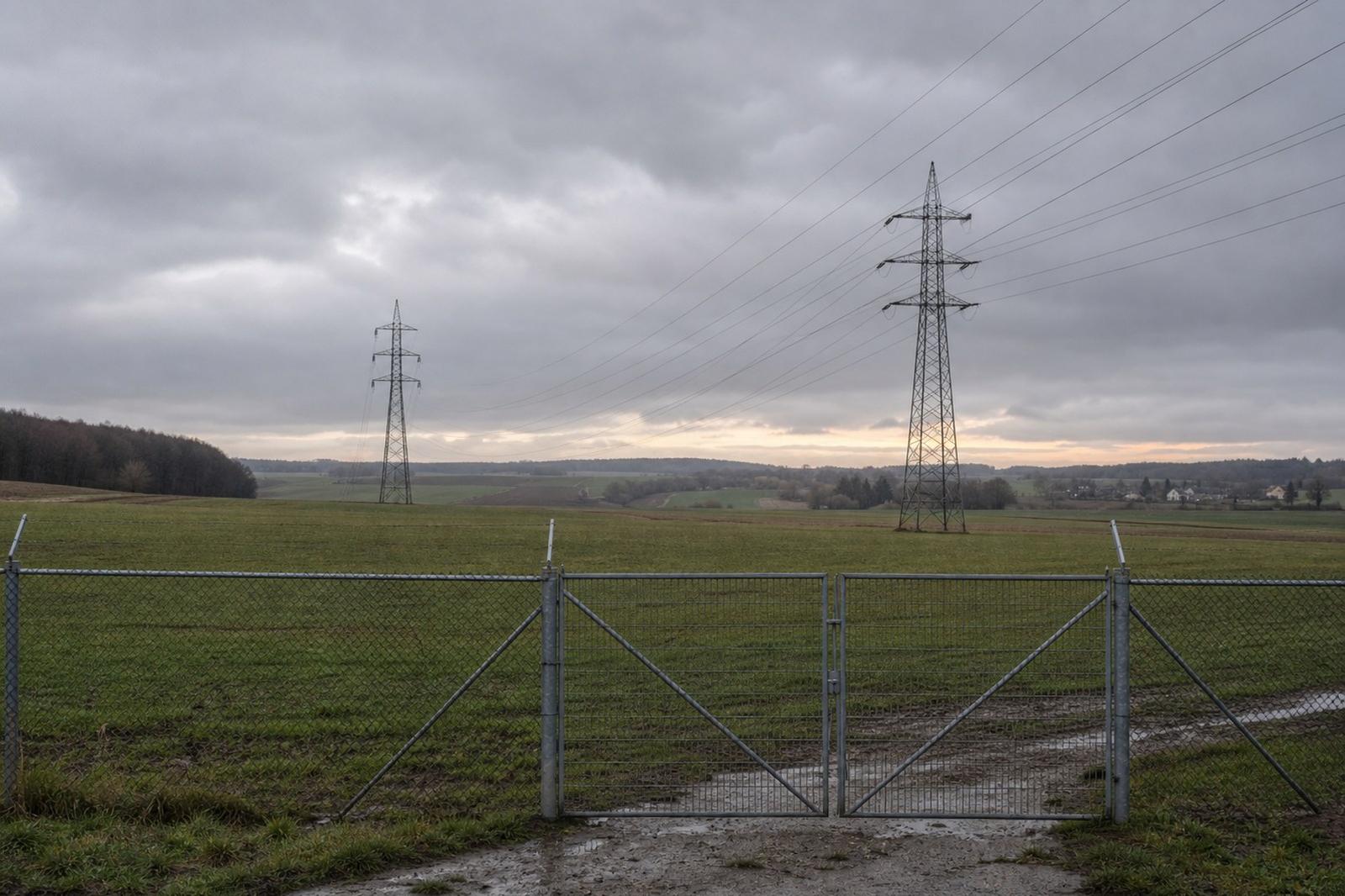 An empty fenced field of farmland near Bissen crossed by high-voltage power lines at dusk, the planned site of Google's data centre.