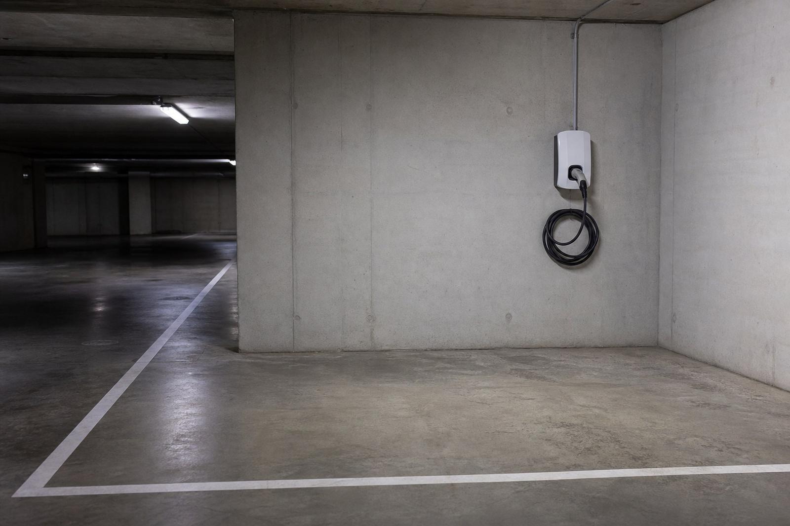 Emplacement de parking vide marqué au sol dans un parking souterrain, à côté d'une borne de recharge murale au câble enroulé.