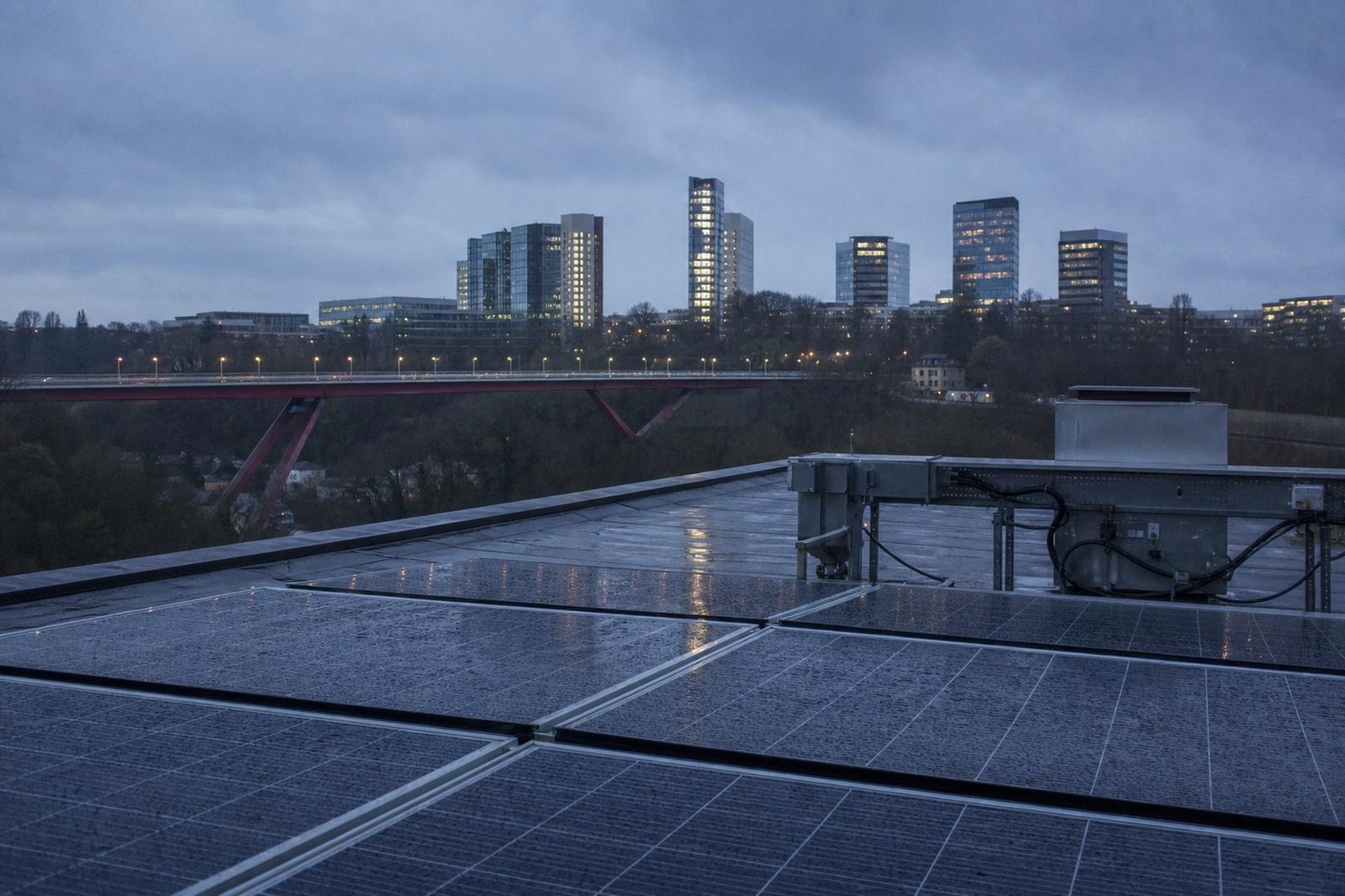 Des panneaux solaires sur un toit de Luxembourg, avec des lignes électriques et le Kirchberg à l’arrière-plan.