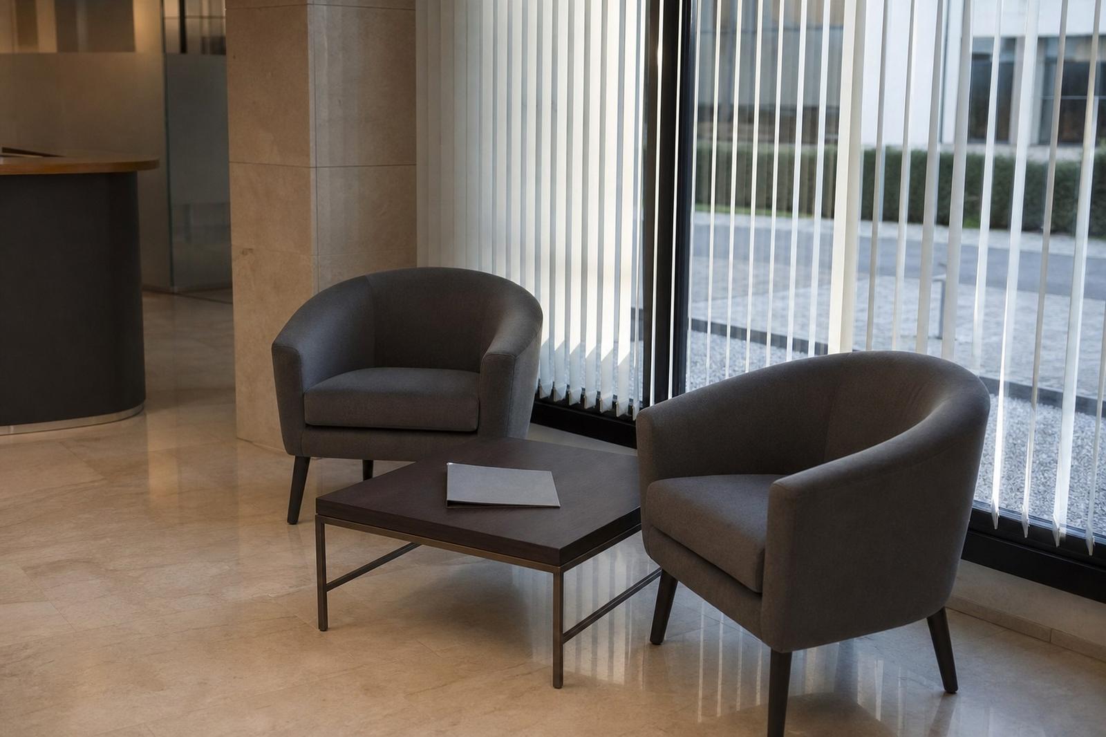 An empty bank branch waiting area with two chairs, a low table holding a closed folder and daylight through half-open blinds.