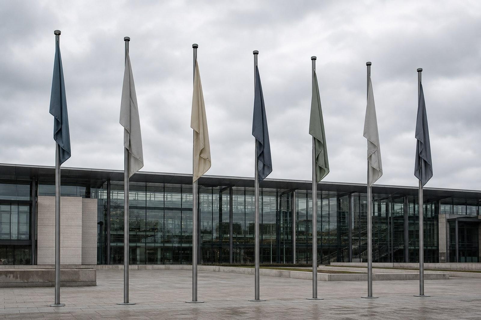 Plain flags hang motionless on a row of poles outside a modern glass office building beneath a grey sky.