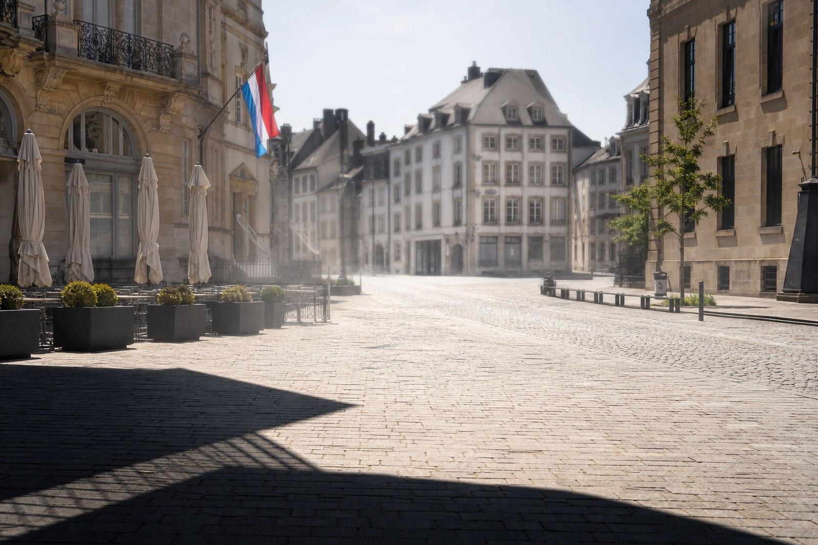 An empty, sun-scorched cobbled square in Luxembourg City at midday, heat haze rising and a national flag hanging limp under a white sky.